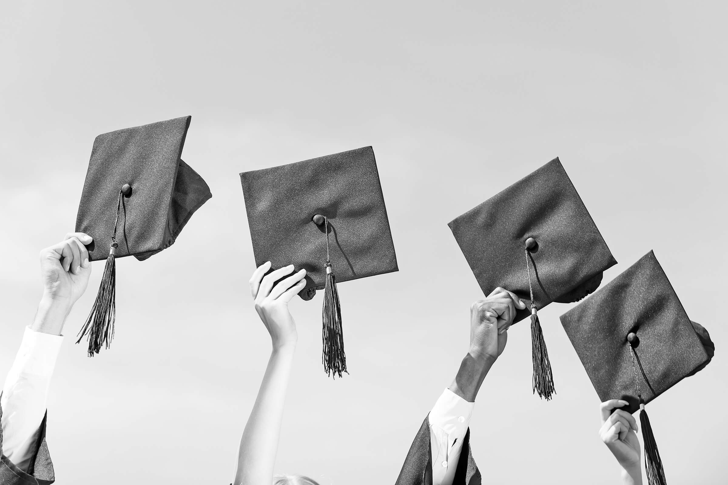 Close-up of four hands holding grad caps against the sky.
